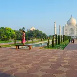 Ein Mann fotografiert ein Mädchen, das auf einer Bank vor dem Taj Mahal steht. Die Anlage des Taj Mahal Land ist mit Bäumen und einem Wasserbecken geschmückt.