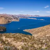 Blick auf den Titicacasee, wo die Anden auf das tiefblaue Wasser treffen. Terrassierte Hänge und eine kleine Insel mit Bäumen zeugen von der Schönheit dieser Region.