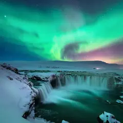 Island reisezeit: Polarlichter tanzen über einem schneebedeckten Wasserfall. Ein magischer Anblick!