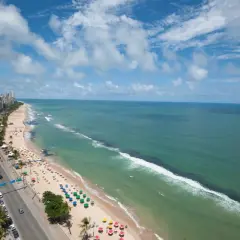 Blick auf den Strand von Recife, Brasilien, mit Palmen, bunten Sonnenschirmen und dem türkisfarbenen Meer unter einem blauen Himmel mit Wolken.