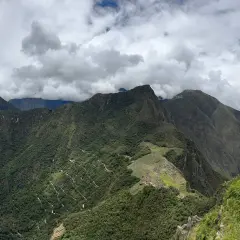 Blick auf Machu Picchu und Huayna Picchu. Eine Frau steht auf einem Felsvorsprung und blickt auf die grüne Berglandschaft.