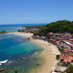 Blick auf die Küste von Morro de São Paulo mit türkisfarbenem Wasser, Sandstrand und bunten Häusern, die sich an einen grünen Hügel schmiegen.