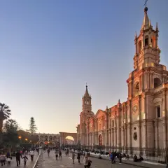 Abendstimmung in Arequipa, Peru. Die Sonne beleuchtet die Kathedrale und die umliegenden Gebäude, während Menschen auf dem Platz spazieren. Die Peru Berge sind in der Ferne zu erahnen.