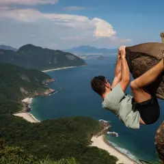 Mann hängt an einem Felsen über dem Meer an der Pedra do Telégrafo. Grüne Hügel und Strände erstrecken sich in der Ferne.