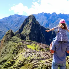 Ein Vater trägt seine Tochter auf den Schultern vor dem majestätischen Machu Picchu Berg. Die antike Stadt liegt eingebettet in die grüne Berglandschaft.