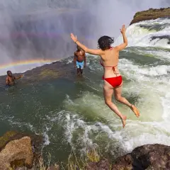 Mutige Frau springt in den Devils Pool am Rand der Victoriafälle, ein Regenbogen bildet sich im Sprühnebel.