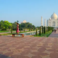 Ein Mann fotografiert ein Mädchen, das auf einer Bank vor dem Taj Mahal steht. Die Anlage des Taj Mahal Land ist mit Bäumen und einem Wasserbecken geschmückt.