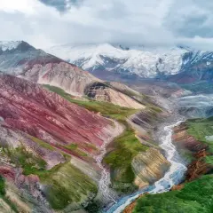 Spektakuläre Kirgisistan Landschaft mit bunten Bergen, einem Fluss und schneebedeckten Gipfeln unter dramatischem Himmel.
