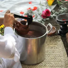 Vorbereitung von Ayahuasca: Eine Person schöpft mit einer Kelle eine braune Flüssigkeit in einen Tonkrug. Blumen und Holz liegen daneben.