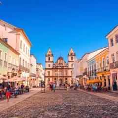 Bunte Kolonialhäuser säumen eine Kopfsteinpflasterstraße in Salvador de Bahia. Menschen flanieren, genießen die Sonne und besuchen Geschäfte.