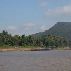 Ein breiter, träger Fluss in Laos, gesäumt von üppiger Vegetation und einem bewaldeten Hügel. Ein langes, blaues Boot liegt am Ufer.