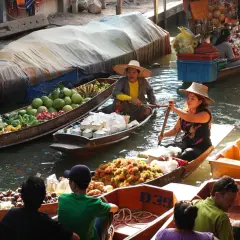 Bunte Boote voller Früchte und Kokosnüsse auf einem schwimmenden Markt in Thailand. Händlerinnen rudern geschickt durch das Wasser.