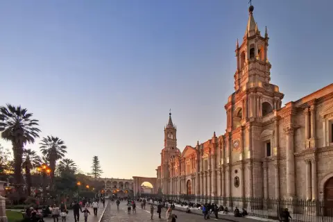 Abendstimmung in Arequipa, Peru. Die Sonne beleuchtet die Kathedrale und die umliegenden Gebäude, während Menschen auf dem Platz spazieren. Die Peru Berge sind in der Ferne zu erahnen.