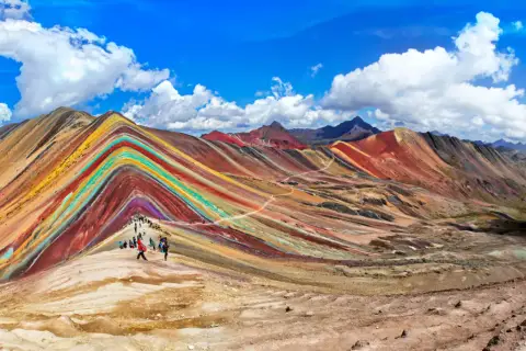 Regenbogenberg in Peru im September. Wanderer bestaunen die bunten Mineralstreifen der Berge unter blauem Himmel.
