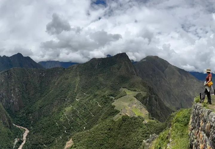 Blick auf Machu Picchu und Huayna Picchu. Eine Frau steht auf einem Felsvorsprung und blickt auf die grüne Berglandschaft.