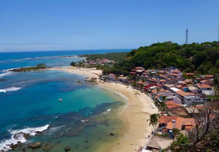 Blick auf die Küste von Morro de São Paulo mit türkisfarbenem Wasser, Sandstrand und bunten Häusern, die sich an einen grünen Hügel schmiegen.