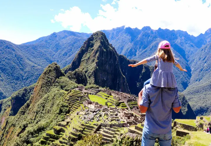 Ein Vater trägt seine Tochter auf den Schultern vor dem majestätischen Machu Picchu Berg. Die antike Stadt liegt eingebettet in die grüne Berglandschaft.
