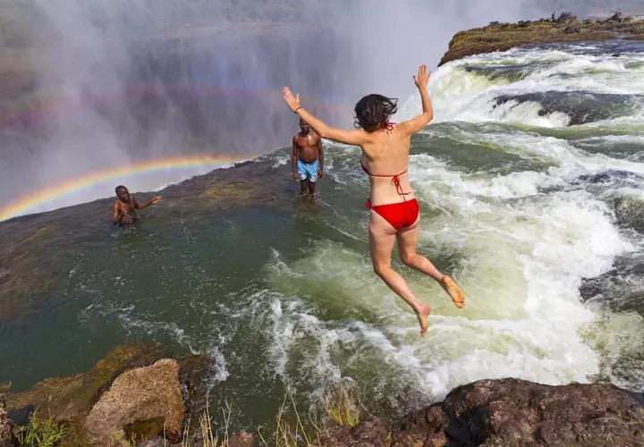 Mutige Frau springt in den Devils Pool am Rand der Victoriafälle, ein Regenbogen bildet sich im Sprühnebel.
