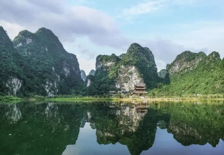 Grüne Berge spiegeln sich im ruhigen Wasser. Ein Tempel schmiegt sich an den Hang. Perfekt für die beste Reisezeit Vietnam.
