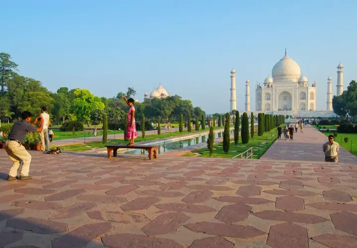 Ein Mann fotografiert ein Mädchen, das auf einer Bank vor dem Taj Mahal steht. Die Anlage des Taj Mahal Land ist mit Bäumen und einem Wasserbecken geschmückt.