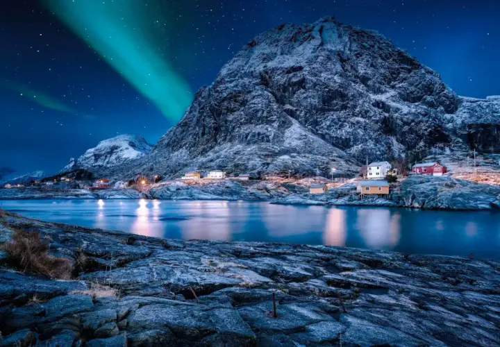 Nordkap Polarlichter tanzen über verschneite Berge und kleine Häuser am Fjord. Ein magischer Anblick!