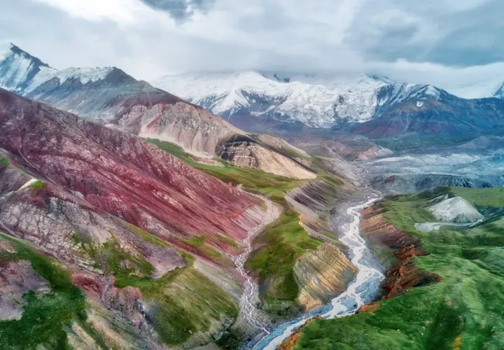 Spektakuläre Kirgisistan Landschaft mit bunten Bergen, einem Fluss und schneebedeckten Gipfeln unter dramatischem Himmel.