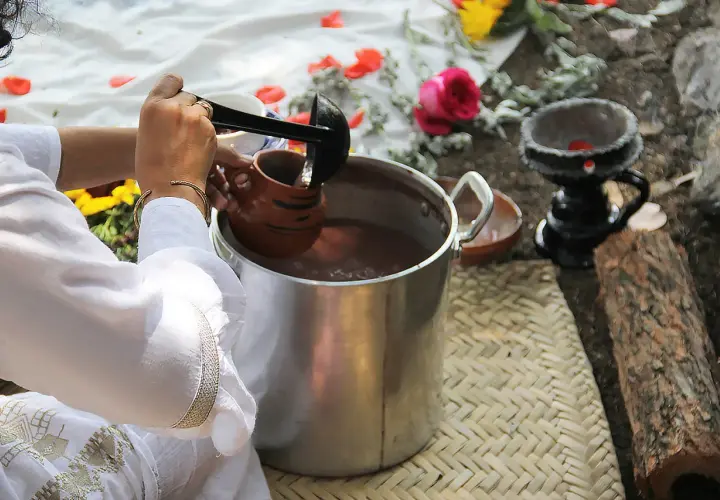 Vorbereitung von Ayahuasca: Eine Person schöpft mit einer Kelle eine braune Flüssigkeit in einen Tonkrug. Blumen und Holz liegen daneben.