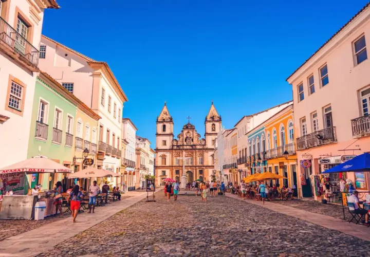 Bunte Kolonialhäuser säumen eine Kopfsteinpflasterstraße in Salvador de Bahia. Menschen flanieren, genießen die Sonne und besuchen Geschäfte.