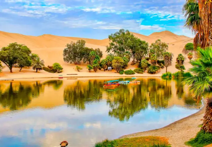 Oase Huacachina bei Ica, Peru, mit Sanddünen, Palmen und einem See voller bunter Boote.