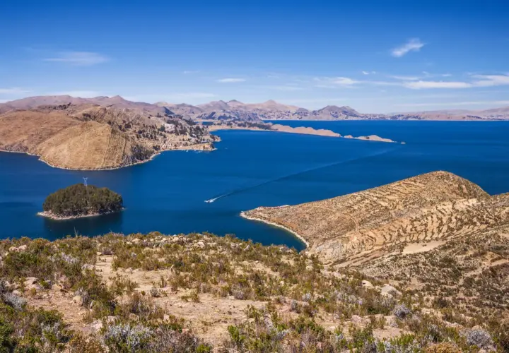 Blick auf den Titicacasee, wo die Anden auf das tiefblaue Wasser treffen. Terrassierte Hänge und eine kleine Insel mit Bäumen zeugen von der Schönheit dieser Region.
