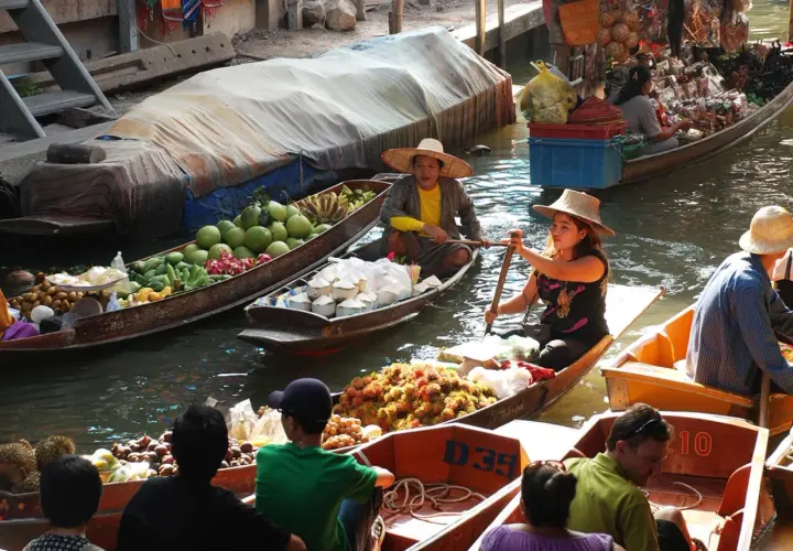 Bunte Boote voller Früchte und Kokosnüsse auf einem schwimmenden Markt in Thailand. Händlerinnen rudern geschickt durch das Wasser.