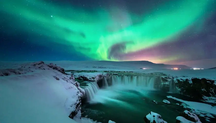 Island reisezeit: Polarlichter tanzen über einem schneebedeckten Wasserfall. Ein magischer Anblick!