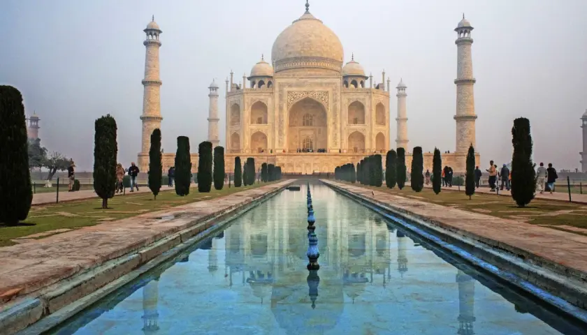 Das Taj Mahal, eine Moschee, spiegelt sich im Wasser. Besucher spazieren durch den Garten.