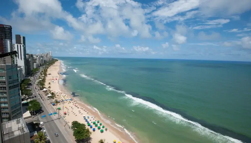 Blick auf den Strand von Recife, Brasilien, mit Palmen, bunten Sonnenschirmen und dem türkisfarbenen Meer unter einem blauen Himmel mit Wolken.