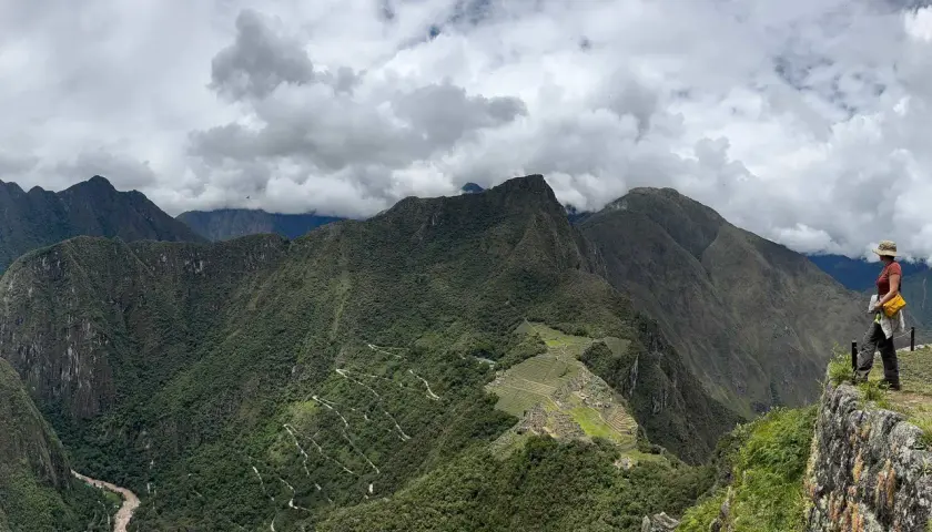 Blick auf Machu Picchu und Huayna Picchu. Eine Frau steht auf einem Felsvorsprung und blickt auf die grüne Berglandschaft.