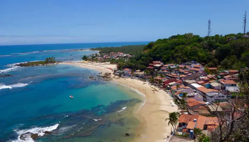Blick auf die Küste von Morro de São Paulo mit türkisfarbenem Wasser, Sandstrand und bunten Häusern, die sich an einen grünen Hügel schmiegen.