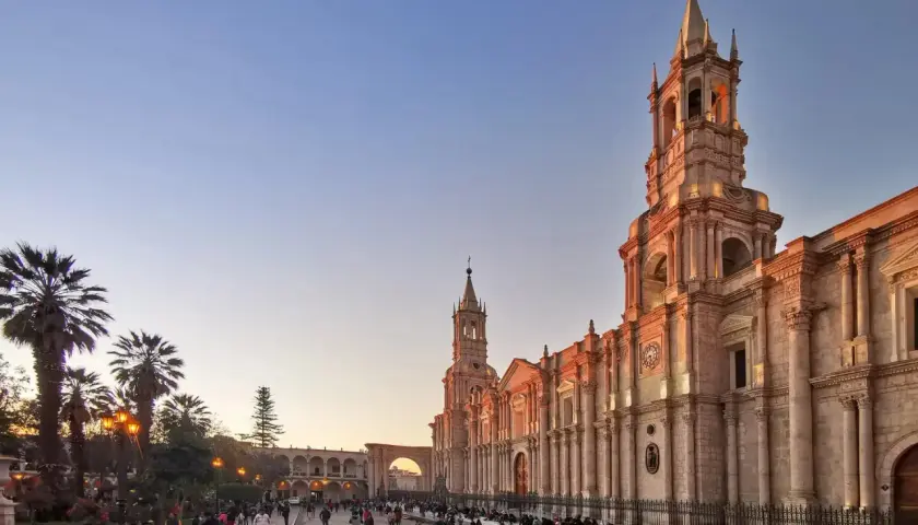 Abendstimmung in Arequipa, Peru. Die Sonne beleuchtet die Kathedrale und die umliegenden Gebäude, während Menschen auf dem Platz spazieren. Die Peru Berge sind in der Ferne zu erahnen.