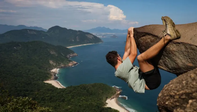 Mann hängt an einem Felsen über dem Meer an der Pedra do Telégrafo. Grüne Hügel und Strände erstrecken sich in der Ferne.