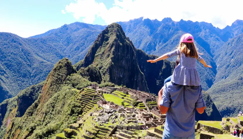 Ein Vater trägt seine Tochter auf den Schultern vor dem majestätischen Machu Picchu Berg. Die antike Stadt liegt eingebettet in die grüne Berglandschaft.