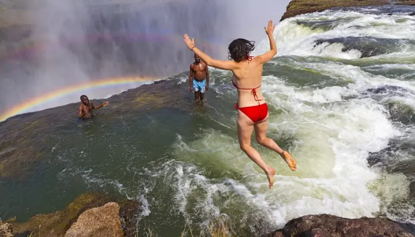 Mutige Frau springt in den Devils Pool am Rand der Victoriafälle, ein Regenbogen bildet sich im Sprühnebel.