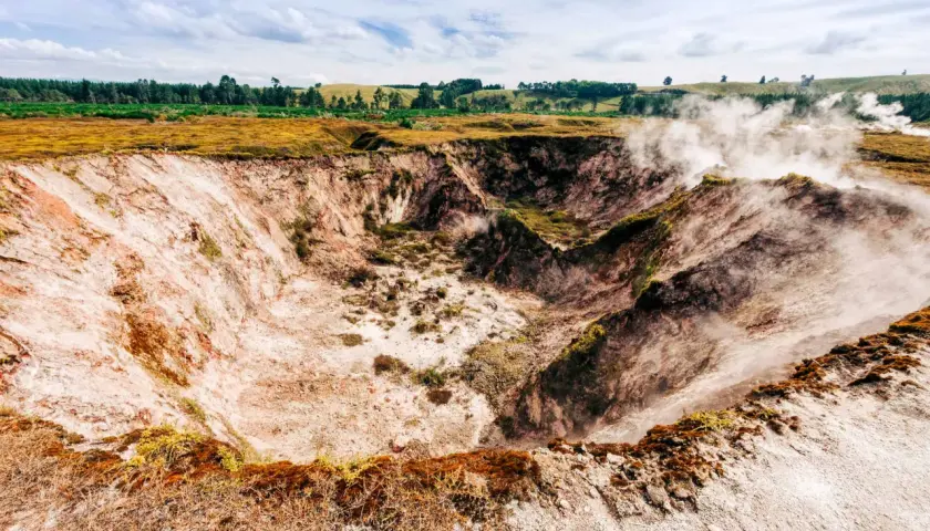 Dampf steigt aus einem Krater in Taupō, Neuseeland. Die Erde ist rötlich-weiß, mit grüner Vegetation am Rand.