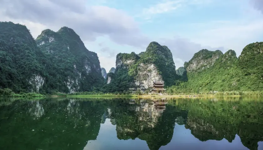Grüne Berge spiegeln sich im ruhigen Wasser. Ein Tempel schmiegt sich an den Hang. Perfekt für die beste Reisezeit Vietnam.