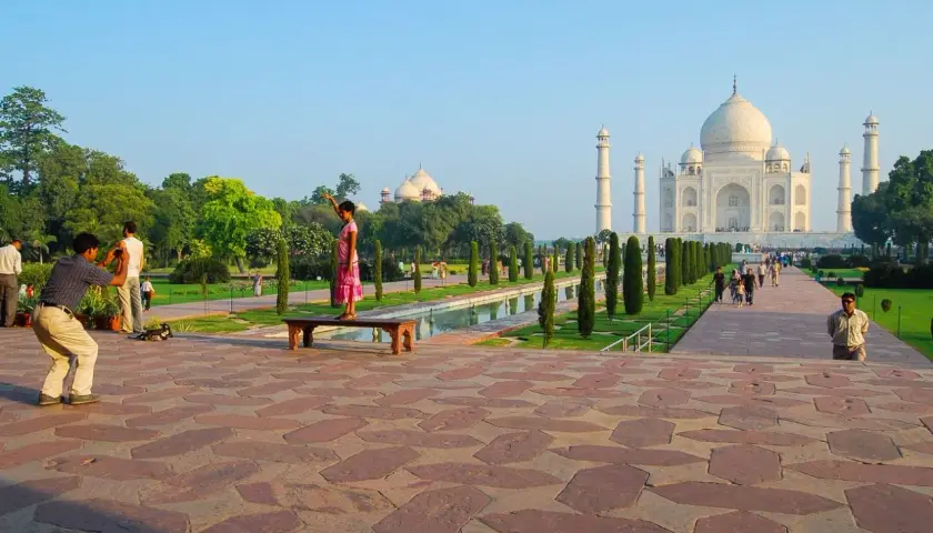 Ein Mann fotografiert ein Mädchen, das auf einer Bank vor dem Taj Mahal steht. Die Anlage des Taj Mahal Land ist mit Bäumen und einem Wasserbecken geschmückt.