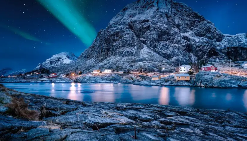 Nordkap Polarlichter tanzen über verschneite Berge und kleine Häuser am Fjord. Ein magischer Anblick!