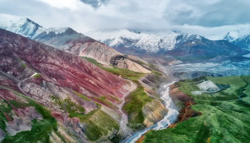 Spektakuläre Kirgisistan Landschaft mit bunten Bergen, einem Fluss und schneebedeckten Gipfeln unter dramatischem Himmel.
