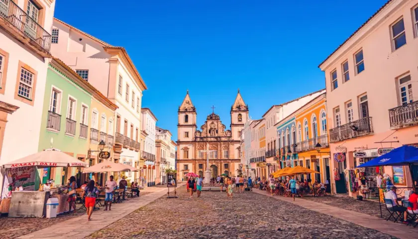 Bunte Kolonialhäuser säumen eine Kopfsteinpflasterstraße in Salvador de Bahia. Menschen flanieren, genießen die Sonne und besuchen Geschäfte.