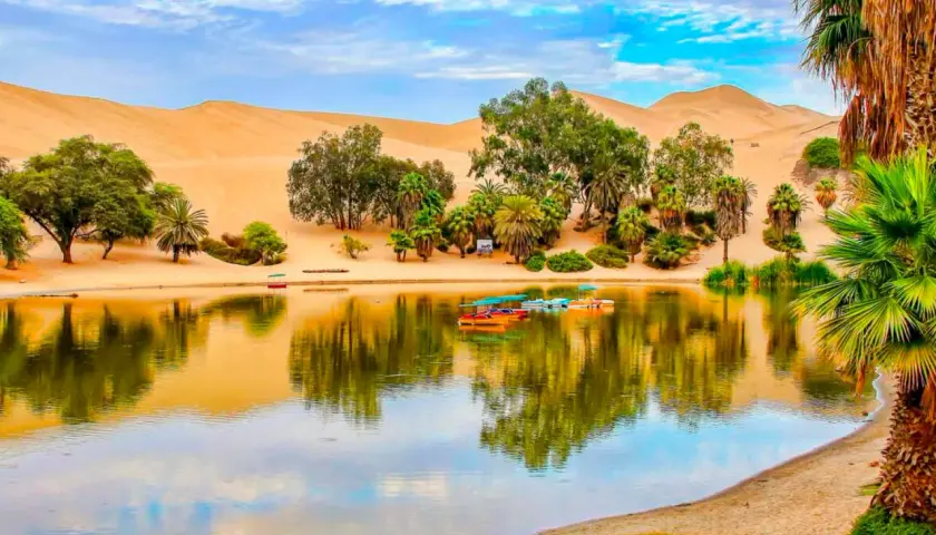 Oase Huacachina bei Ica, Peru, mit Sanddünen, Palmen und einem See voller bunter Boote.