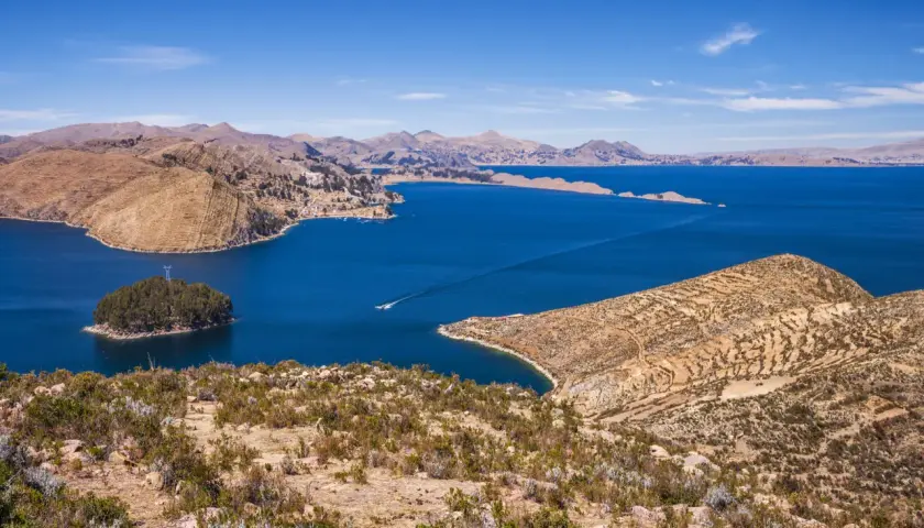 Blick auf den Titicacasee, wo die Anden auf das tiefblaue Wasser treffen. Terrassierte Hänge und eine kleine Insel mit Bäumen zeugen von der Schönheit dieser Region.