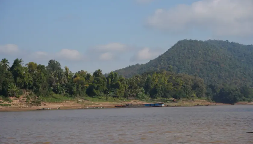 Ein breiter, träger Fluss in Laos, gesäumt von üppiger Vegetation und einem bewaldeten Hügel. Ein langes, blaues Boot liegt am Ufer.
