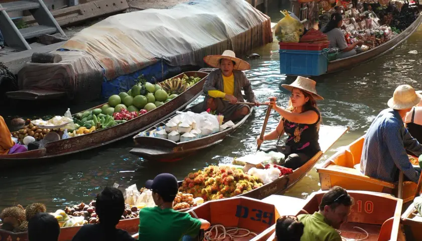 Bunte Boote voller Früchte und Kokosnüsse auf einem schwimmenden Markt in Thailand. Händlerinnen rudern geschickt durch das Wasser.
